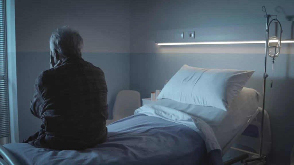 A photo of the back of an older gentleman sitting on the edge of a bed and looking out the window. He is at a nursing home or other long-term care facility.  