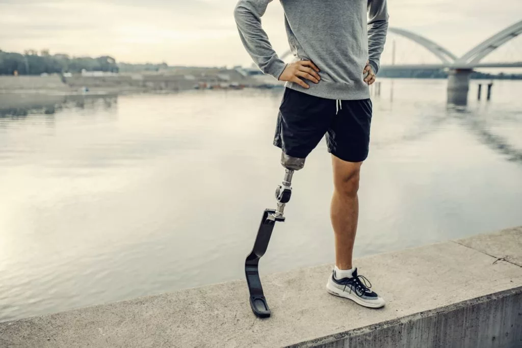 A photo of a man with a prosthetic leg standing on a ledge next to a body of water. He is wearing shorts and an athletic top. 