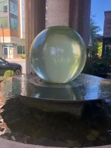 A photo of a large fountain that looks like a green marble. The water is flowing down the round ball into a flat fountain before flowing down the edges into a bunch of rocks. 